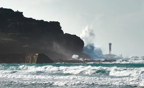 Longships cliffs waves This landscape photograph captures dramatic waves crashing against the Longships cliffs on the coast of Cornwall, United Kingdom, during the early afternoon in winter. The rugged cliffs of Sennen Cove rise prominently on the left of the image, while the powerful sea surges ashore, sending plumes of spray into the air. In the distance, the Longships Lighthouse stands as a landmark, watching over the tumultuous waters of this section of the Cornish coast. The winter light highlights the natural features of the cliffs, the dynamic motion of the sea, and the enduring presence of the lighthouse, all characteristic of this wild part of nature in the United Kingdom.